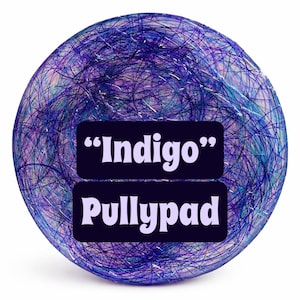 May include: A circular, translucent pad featuring a dense, chaotic web of fine purple, blue, and silver fibers embedded within the material. Centered on the surface are two black rectangular labels with white text reading "Indigo" and "Pullypad".