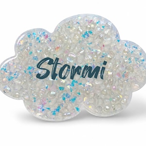 May include: A cloud-shaped decorative object filled with clear, iridescent, and blue-tinted beads and small reflective flakes. The word Stormi is printed in dark blue script across the center of the clear, rounded, cloud-like structure.