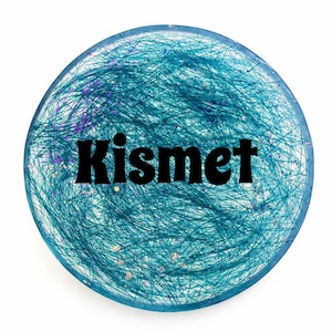 May include: A round, teal-colored button with the word "Kismet" in bold black letters. The button's surface features a textured design of teal fibers and flecks of purple and gold glitter.