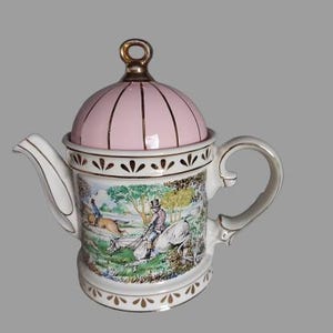 Pode incluir: Bule de porcelana vintage com tampa rosa e detalhes dourados. O bule apresenta uma cena de caça. A alça e o bico são brancos com acabamentos dourados, e a tampa tem listras douradas verticais.