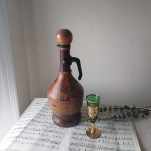 May include: A vintage decanter with a wooden stopper and a handle, featuring a map design. Beside it is a green and gold ornate glass. The items are on a sheet of music.