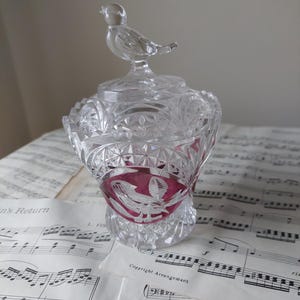 May include: A clear glass jar with a lid topped by a bird figurine. The jar has a cut-glass design and a band of red glass with a white etched design. The jar is sitting on top of sheet music.