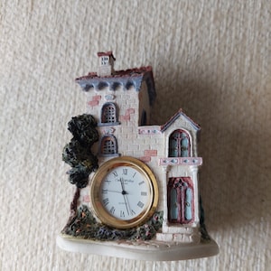 May include: A miniature house-shaped clock with a white clock face and black Roman numerals. The house is made of brick and stone, with a red roof and a small tree. The clock face has gold-coloured hands.