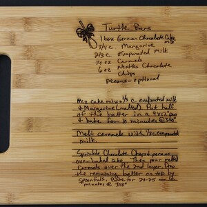 May include: A close-up of a wooden cutting board with a handwritten recipe for Turtle Bars. The recipe includes ingredients like German chocolate cake mix, margarine, evaporated milk, caramels, and chocolate chips. The recipe also includes instructions for baking the bars.