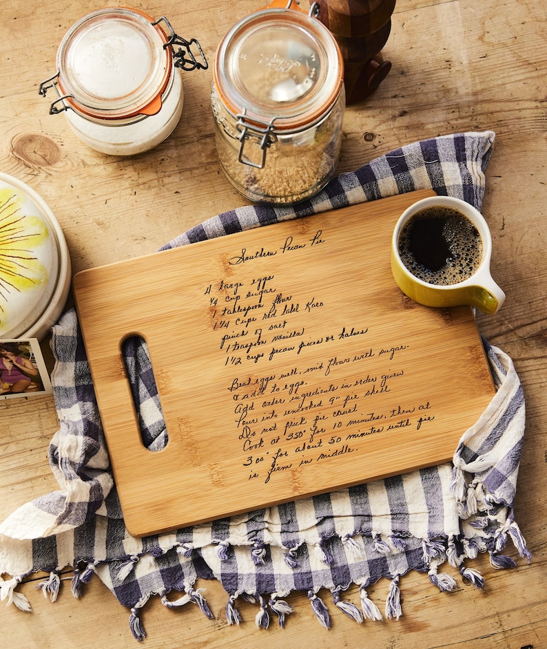 May include: A wooden cutting board with a handwritten recipe for Southern Pecan Pie. The recipe includes ingredients like sugar, flour, pecans, and eggs. The board is on a checkered blue and white cloth.