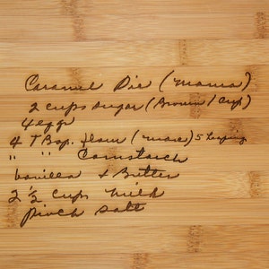 May include: A handwritten recipe for Caramel Pie on a wooden cutting board. The recipe includes 2 cups of sugar, 4 eggs, 4 tablespoons of flour, cornstarch, vanilla, butter, 2 1/2 cups of milk, and a pinch of salt.