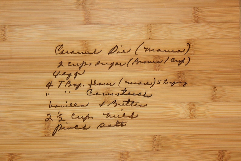 May include: A handwritten recipe for Caramel Pie on a wooden cutting board. The recipe includes 2 cups of sugar, 4 eggs, 4 tablespoons of flour, cornstarch, vanilla, butter, 2 1/2 cups of milk, and a pinch of salt.