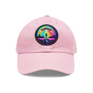 Może przedstawiać: Różowa czapka z daszkiem z haftowanym czarno-białym naszywką przedstawiającą osobę stojącą w lesie z tęczą i UFO nad nią.