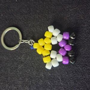 Peut inclure: Un porte-cl&eacute;s avec un drapeau de la fiert&eacute; non binaire. Le drapeau est compos&eacute; de perles jaunes, blanches, violettes et noires. Le porte-cl&eacute;s a un anneau et une cha&icirc;ne en m&eacute;tal argent&eacute;.