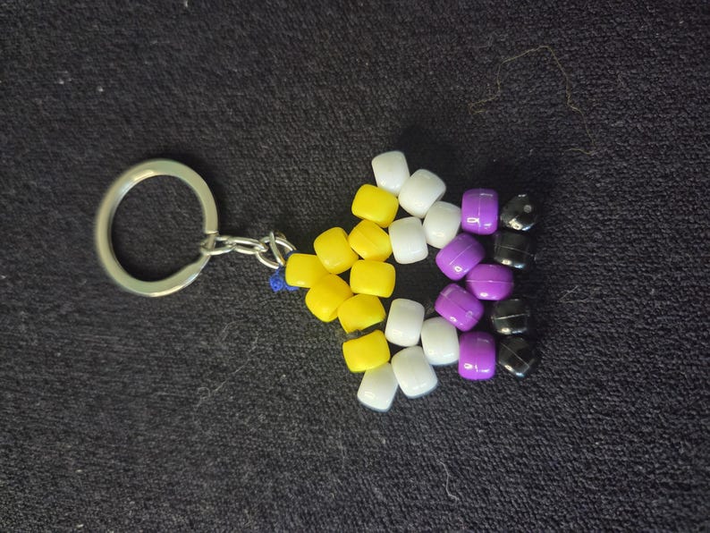 Peut inclure: Un porte-cl&eacute;s avec un drapeau de la fiert&eacute; non binaire. Le drapeau est compos&eacute; de perles jaunes, blanches, violettes et noires. Le porte-cl&eacute;s a un anneau et une cha&icirc;ne en m&eacute;tal argent&eacute;.