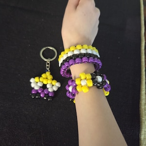 May include: Non-binary pride bracelets and a keychain. The bracelets are made of yellow, white, black, and purple beads. The keychain is a star shape with the same color scheme. The items are on a black surface.