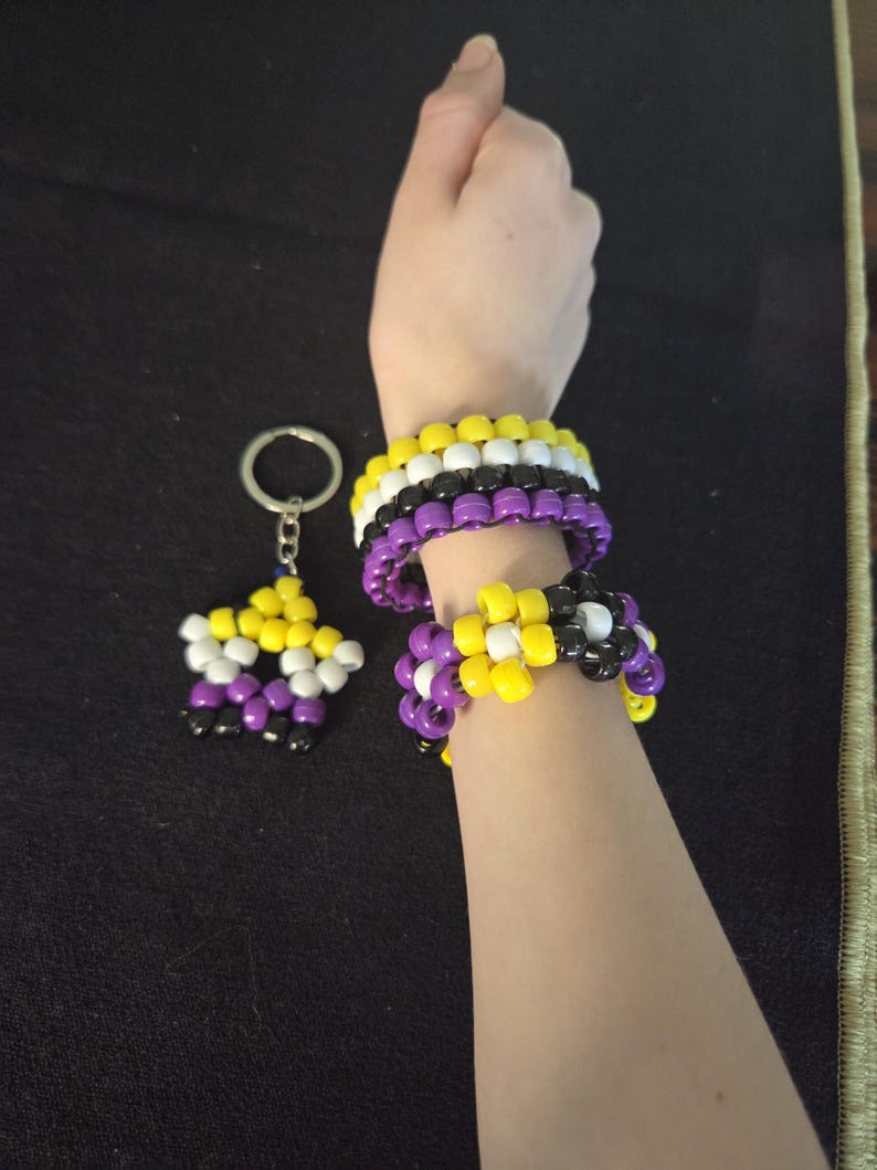 Peut inclure: Bracelets et porte-cl&eacute;s aux couleurs de la fiert&eacute; non-binaire. Les bracelets sont compos&eacute;s de perles jaunes, blanches, noires et violettes. Le porte-cl&eacute;s est en forme d'&eacute;toile et reprend les m&ecirc;mes couleurs. Sur fond noir.