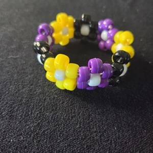 Peut inclure: Un bracelet de perles aux couleurs du drapeau de la fiert&eacute; non binaire : jaune, violet, noir et blanc. Le bracelet est compos&eacute; de petites perles rondes assembl&eacute;es pour former une forme circulaire. Les perles sont dispos&eacute;es en motif.