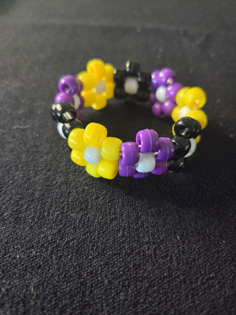 Peut inclure: Un bracelet de perles aux couleurs du drapeau de la fiert&eacute; non binaire : jaune, violet, noir et blanc. Le bracelet est compos&eacute; de petites perles rondes assembl&eacute;es pour former une forme circulaire. Les perles sont dispos&eacute;es en motif.