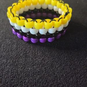 Peut inclure: Un bracelet de perles aux couleurs du drapeau de la fiert&eacute; non binaire : jaune, blanc, noir, violet. Le bracelet est compos&eacute; de petites perles rondes enfil&eacute;es ensemble. Les couleurs sont dispos&eacute;es en rayures horizontales.