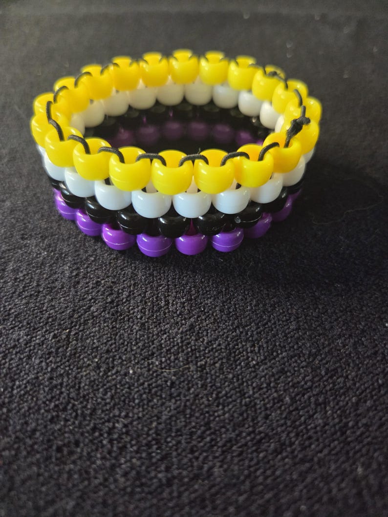 Peut inclure: Un bracelet de perles aux couleurs du drapeau de la fiert&eacute; non binaire : jaune, blanc, noir, violet. Le bracelet est compos&eacute; de petites perles rondes enfil&eacute;es ensemble. Les couleurs sont dispos&eacute;es en rayures horizontales.