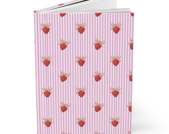 May include: A hardcover journal with a pink and white striped pattern. The cover is adorned with a repeating pattern of red strawberries tied with pink bows. The journal is open, revealing blank white pages.