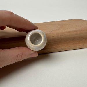 May include: A small, round, ceramic object with a central hole, held by a hand. The object has a light, iridescent glaze with hints of blue and white. The background includes a wooden cutting board and a white surface.