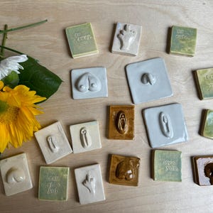 May include: A collection of ceramic tiles with various shapes and designs, including body parts and text. The tiles are in shades of white, brown, and green. A yellow sunflower and other flowers are in the background.