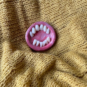 May include: A pink ceramic dish shaped like a mouth with white teeth. The dish is on a yellow knitted fabric.