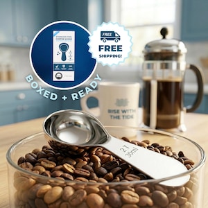 May include: A stainless steel coffee scoop, 2T (30ml), rests on coffee beans in a glass container. The image includes a coffee press, a mug with the text "RISE WITH THE TIDE", and a box with the text "STAINLESS STEEL COFFEE SCOOP".