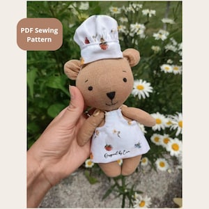 May include: A small brown fabric teddy bear wearing a white chef hat and a white apron with a berry print. The apron features the text Original by Eva. The bear has embroidered black eyes and a nose, and the image includes a PDF Sewing Pattern label.