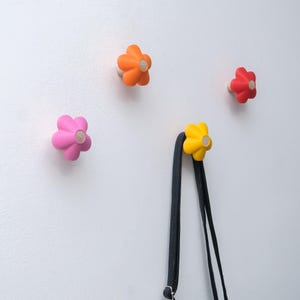 Op de afbeelding: Vier bloemvormige wandhaken in de kleuren roze, oranje, rood en geel. De haken zijn bevestigd aan een witte muur, met een zwarte riem die aan de gele haak hangt. Elke haak heeft een houten midden.