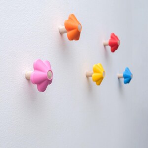 May include: Five wall hooks shaped like stylized flowers, each featuring a light-colored wooden peg base and a vibrant, solid-colored plastic top in orange, red, pink, yellow, and blue, mounted individually on a plain white wall surface.