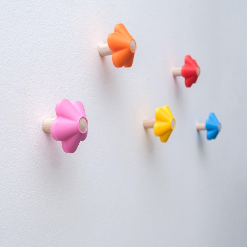 May include: Five wall hooks shaped like stylized flowers, each featuring a light-colored wooden peg base and a vibrant, solid-colored plastic top in orange, red, pink, yellow, and blue, mounted individually on a plain white wall surface.