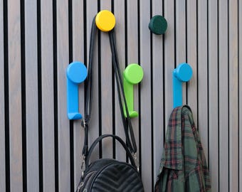 May include: A set of colorful wall-mounted hooks in yellow, blue, and green, featuring a circular top and a vertical arm. A black leather-textured backpack hangs from the yellow hook, and a green and black plaid shirt hangs from the blue hook.
