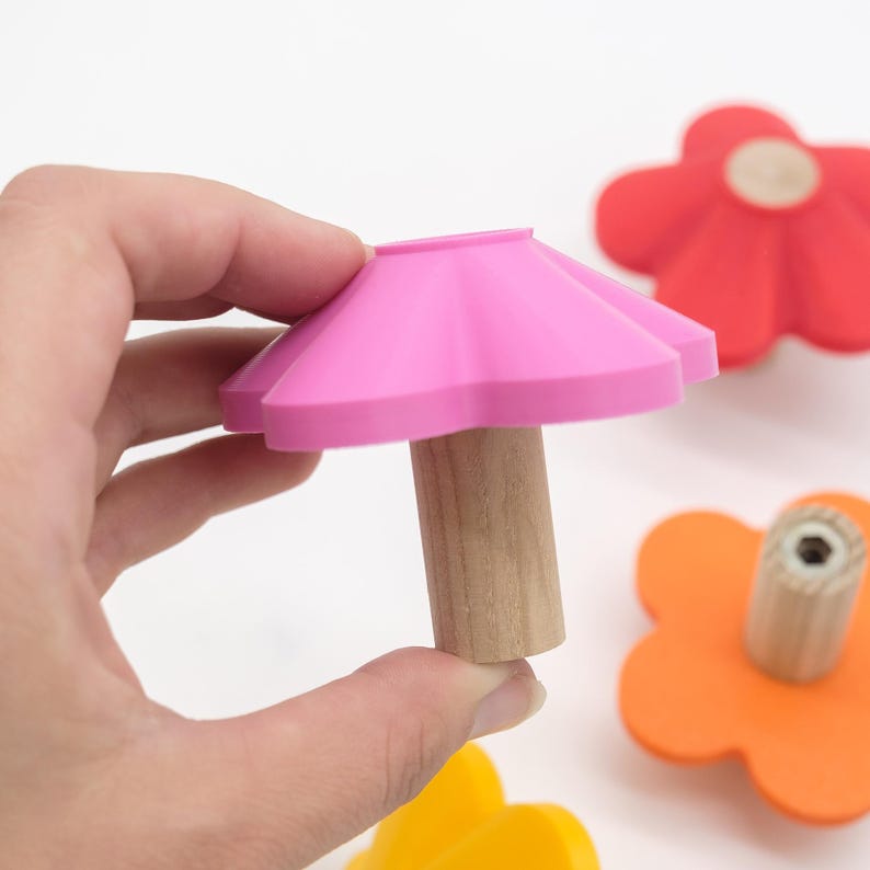 May include: A flower-shaped cabinet knob with a bright pink silicone top and a cylindrical light-colored wooden base. The knob features a smooth, matte finish and a central metal screw hole on the underside for mounting to furniture surfaces.