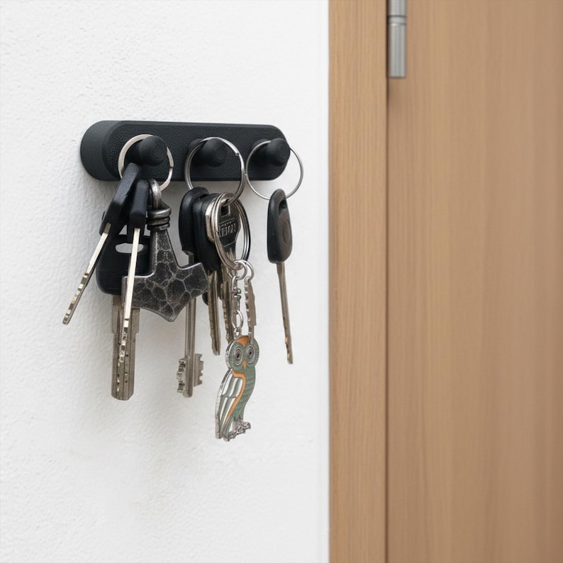 May include: A black magnetic key holder fixed to a white wall, holding various keys. The keyrings include a decorative owl and other metal keys. The holder is near a wooden door.