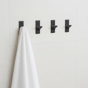 May include: Four black metal wall hooks mounted on a white tiled wall. A white towel hangs from the first hook. The hooks are rectangular with a curved projection for hanging items. Bathroom decor.