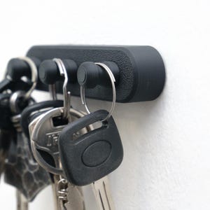 May include: A black key holder with three key hooks. The key holder is mounted on a white wall and holds several keys. The key holder is made of a textured material.