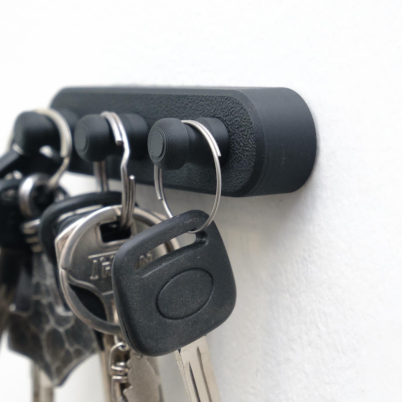 May include: A black key holder with three key hooks. The key holder is mounted on a white wall and holds several keys. The key holder is made of a textured material.