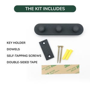 May include: A black key holder with three pegs, a mounting plate, screws, dowels, and double-sided tape. The text "THE KIT INCLUDES" is at the top. The kit includes all the necessary hardware for installation.