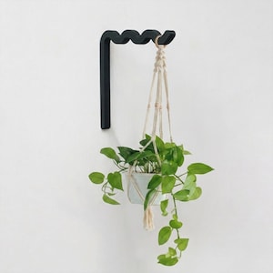 Puede incluir: Un soporte de pared negro para plantas con un borde superior ondulado y serpenteante. Un colgador de macramé de color beige claro cuelga del extremo del soporte, sosteniendo una maceta blanca con una planta pothos de hojas verdes acorazonadas.
