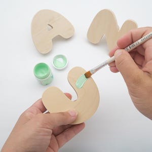 May include: A light-colored wooden letter S being painted with mint green acrylic paint using a small brush. Two additional unpainted wooden letters, A and N, are visible in the background along with a small open container of mint green paint.
