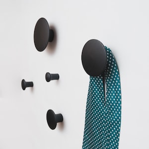 May include: A set of four black wooden wall hooks in various sizes. One hook holds a teal scarf with white polka dots. The hooks are mounted on a white wall, offering a minimalist and modern design.
