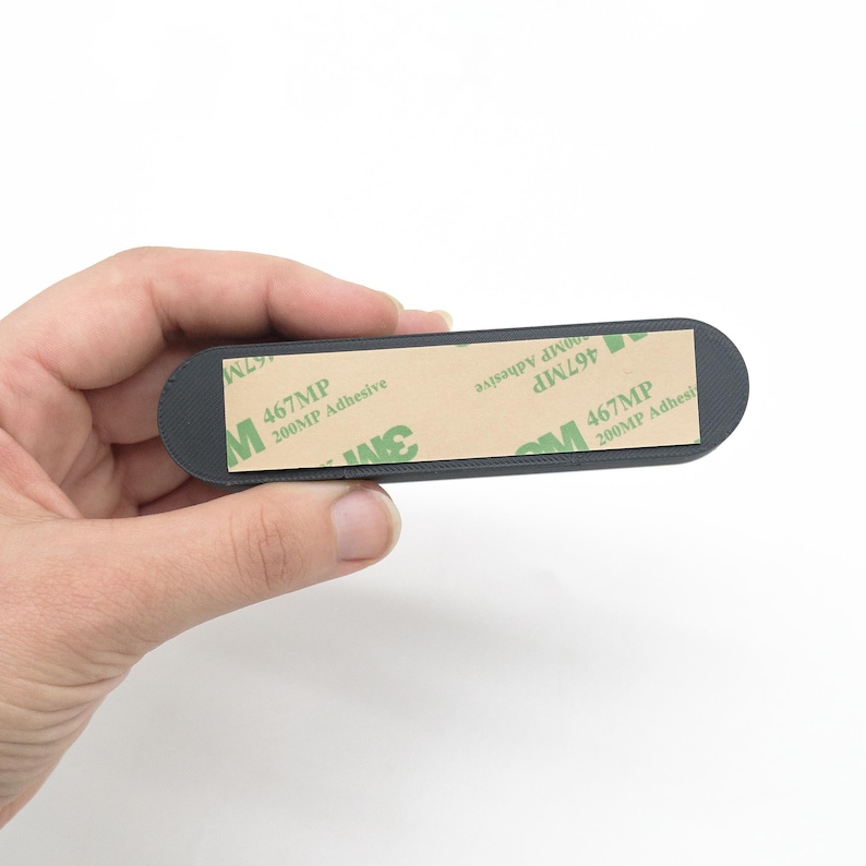 May include: A dark grey, rectangular adhesive mount with rounded ends. The mount has a 3M adhesive strip on the back, with the text "467MP 200MP Adhesive". The mount is held in a hand against a white background.
