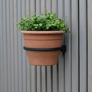 Op de afbeelding: Een terracotta bloempot gevuld met groene planten, gemonteerd op een zwarte metalen beugel. De pot is bevestigd aan een grijze houten schutting, waardoor een eenvoudige maar elegante presentatie voor buitenruimtes ontstaat.
