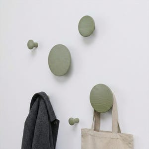 May include: A set of five sage green circular wall hooks of varying sizes mounted on a wall. Two of the hooks are small knobs, while three are larger, flat, disc-shaped hooks. A dark grey coat hangs from one hook and a beige tote bag from another.