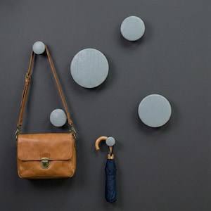 May include: A set of five circular, light gray wall-mounted hooks of varying sizes. A tan leather shoulder bag with a metal clasp hangs from one hook, and a folded dark blue umbrella with a curved wooden handle hangs from another hook.