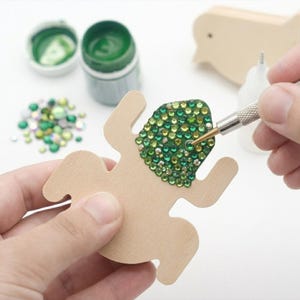 Puede incluir: Un recorte de madera clara con forma de rana que se está decorando con pequeños diamantes de imitación redondos de color verde y amarillo. Se utiliza una herramienta aplicadora de metal para colocar las gemas. Cerca hay un bote de pintura verde y gemas sueltas.