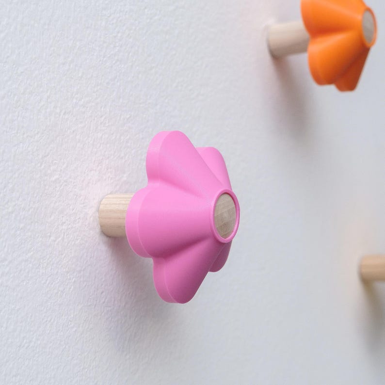May include: A wall-mounted hook featuring a bright pink, five-petaled flower-shaped head made of a smooth material, attached to a light-colored wooden cylindrical base. A second orange flower-shaped hook is visible in the upper right corner.