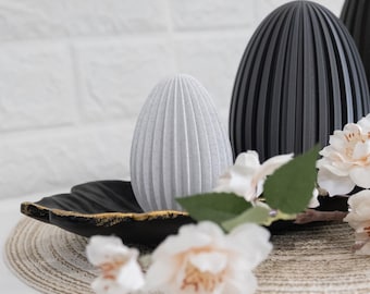 May include: A light gray egg-shaped object with vertical ridges sits next to a larger black ridged egg-shaped object. Both rest on a black leaf-shaped tray with gold-painted edges, accompanied by white artificial flowers.