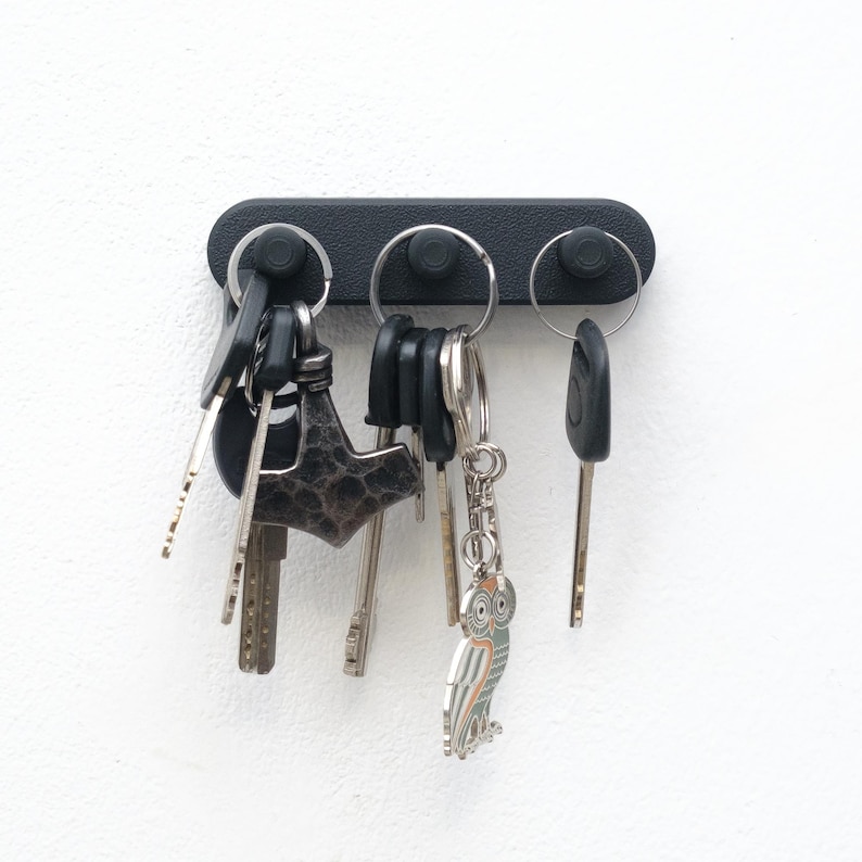 May include: A black key holder with three key rings, each holding multiple keys. The keys are of various shapes and sizes, including a car key and a decorative owl keyring. The key holder is mounted on a white wall.
