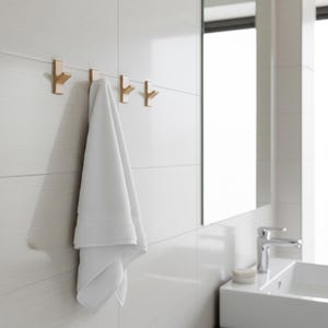 May include: A white towel hangs from a set of three gold-colored wall hooks. The hooks are mounted on a white tiled wall. The bathroom setting includes a mirror, sink, and faucet.