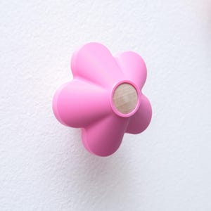 May include: A bright pink wall hook shaped like a five-petaled flower. The center of the flower features a circular, light-colored natural wood insert. The hook has a smooth, matte finish and a rounded, three-dimensional design.