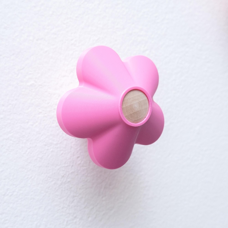 May include: A bright pink wall hook shaped like a five-petaled flower. The center of the flower features a circular, light-colored natural wood insert. The hook has a smooth, matte finish and a rounded, three-dimensional design.